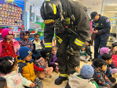 Firefighter demonstrating safety gear to a group of children in a classroom setting.