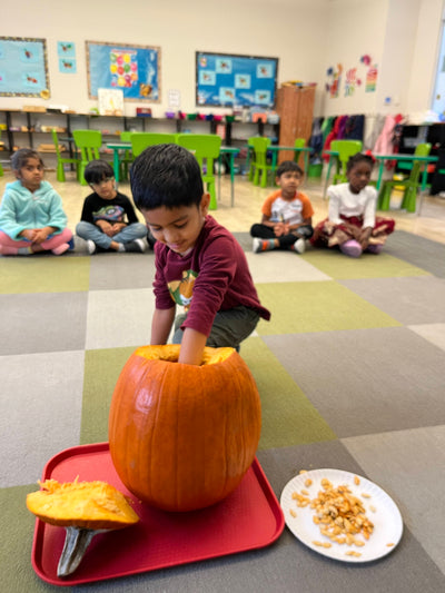Child holding a pumpkin in a classroom setting with other children and educational materials.