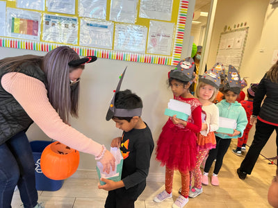 Children in costumes receiving treats from a woman in a classroom setting.