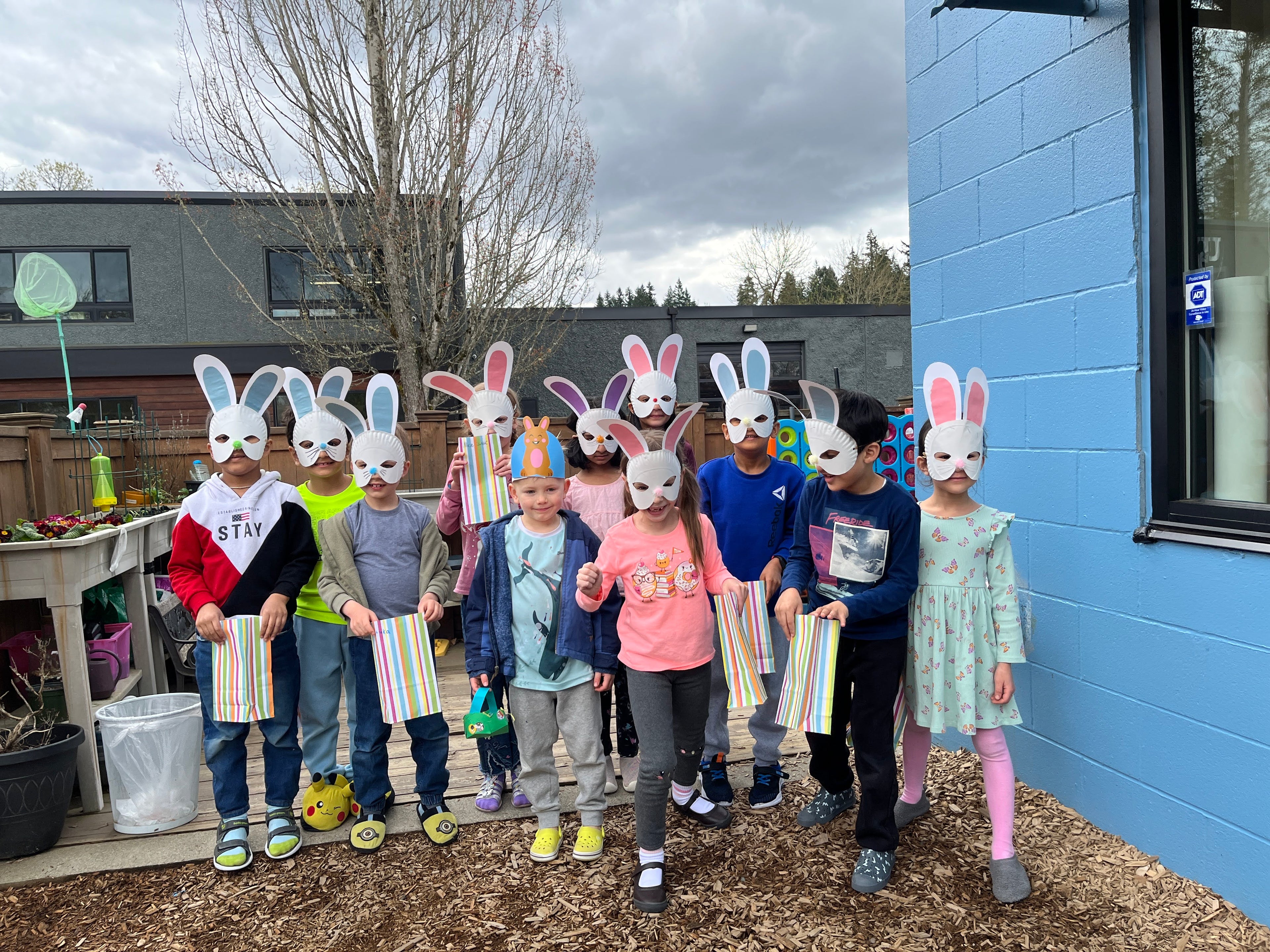 Children wearing bunny masks and holding Easter baskets in front of a blue building.